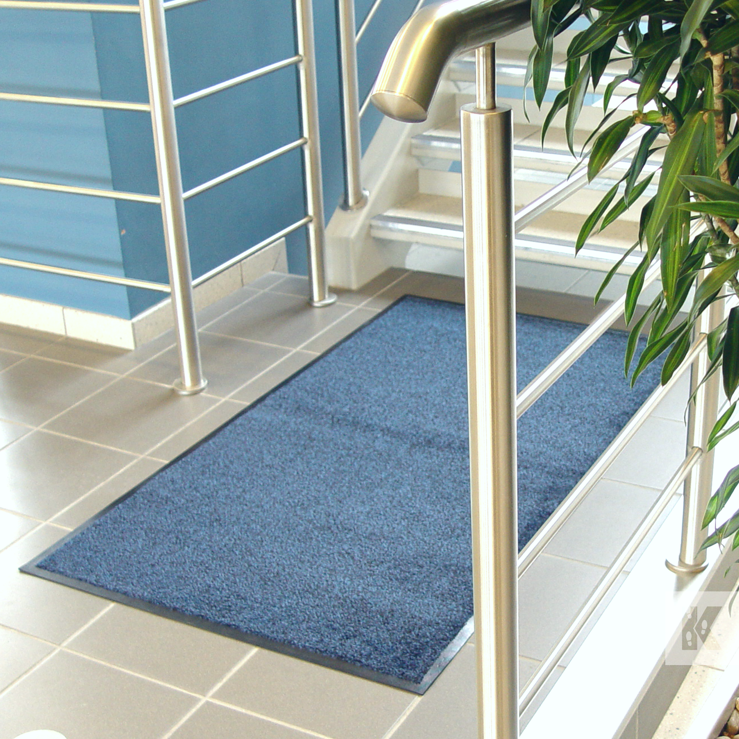 Iron-Horse - A rectangular blue entrance mat made with rubber backing, placed on a tiled floor with a metal handrail and a potted plant in the background.