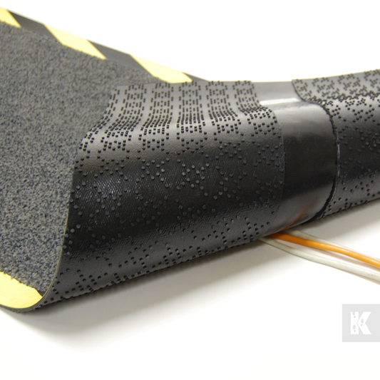 A close-up of a black textured protective sleeve or cable jacket with a perforated dot pattern, showing yellow edges and a small section of exposed orange and white wire, on a white background.