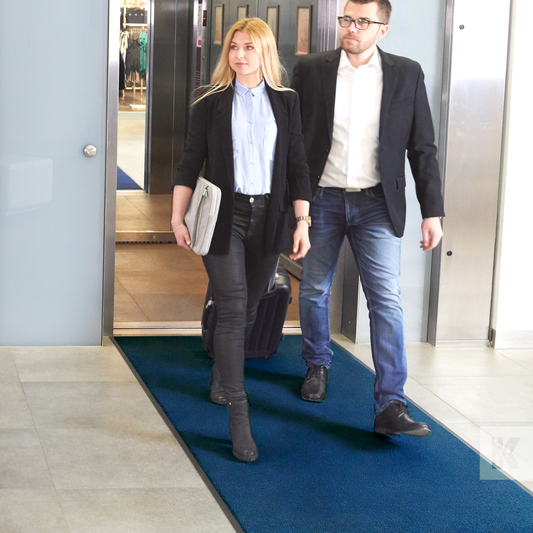 Iron-Horse Mono - Two people walking on a blue rectangular entrance mat placed on a tiled floor inside a building.